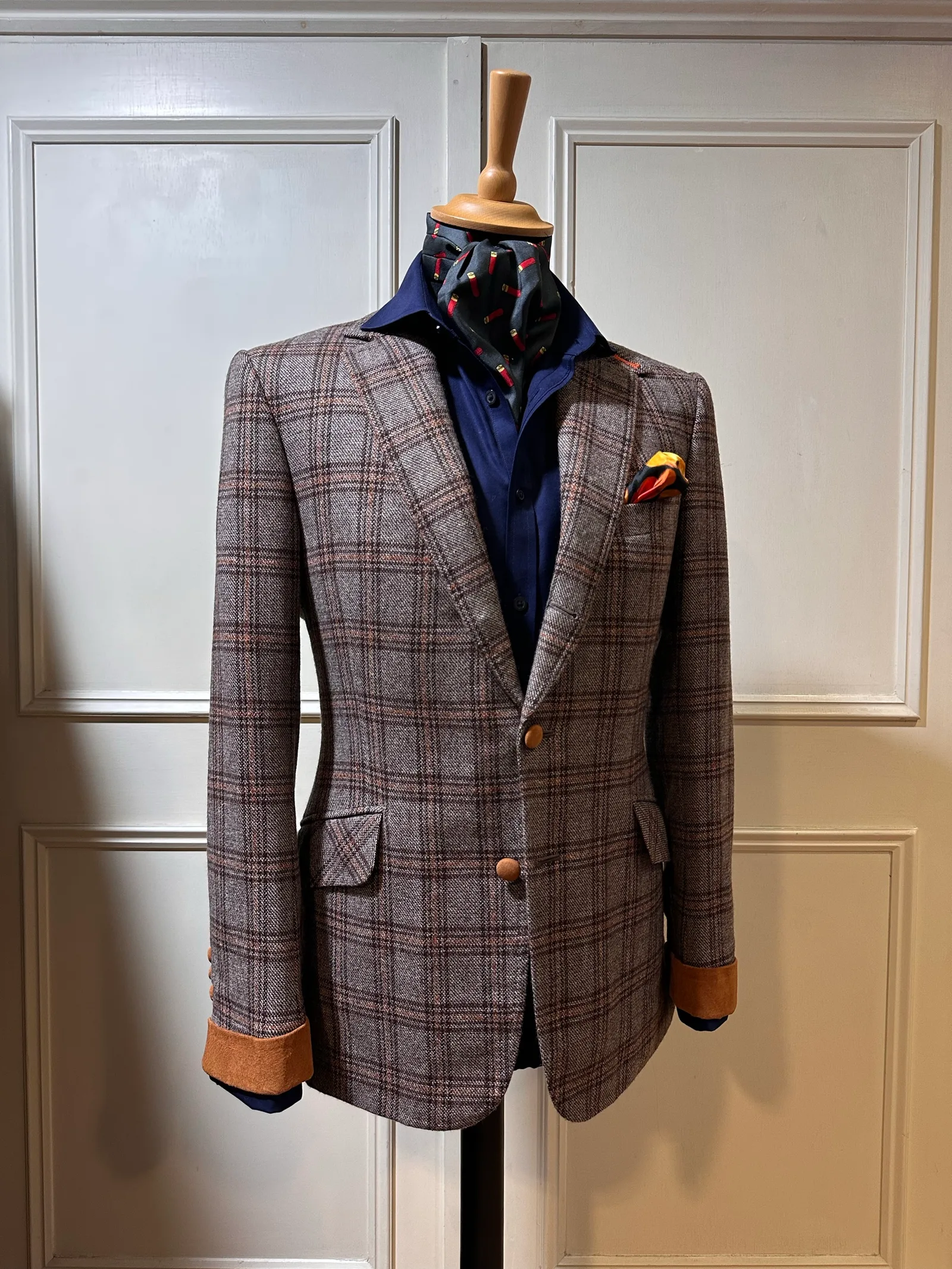 Front mannequin view of a brown Glen-check jacket with suede cuffs, showing notch lapels, three brown horn buttons, shaped waist and tan suede cuffs.