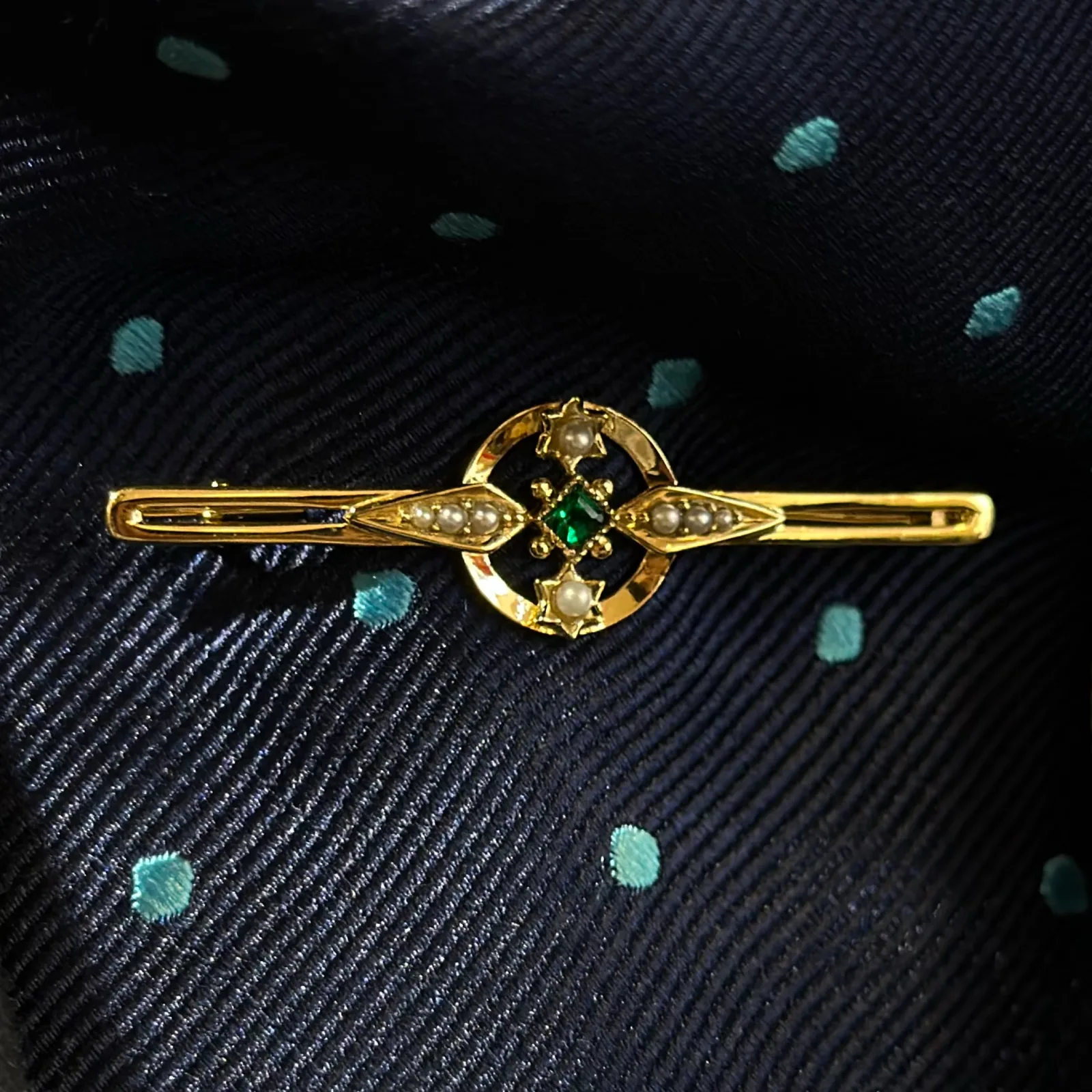Gold tie bar with a square emerald, seed-pearl leaves and an open circular centre resting on navy dotted silk.