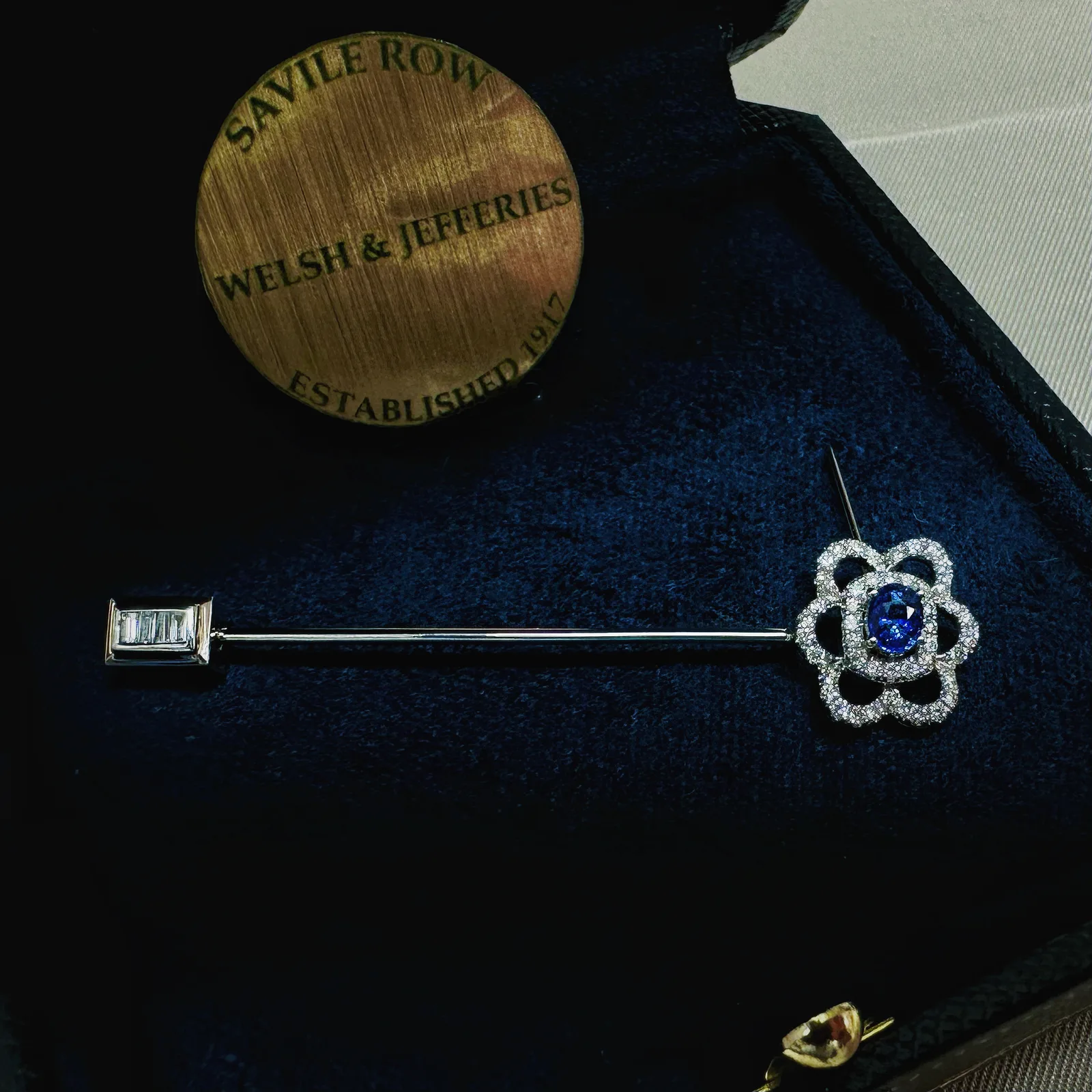 Blue sapphire stick pin encircled by airy diamond petals, with a baguette-set rectangular guard on navy velvet.