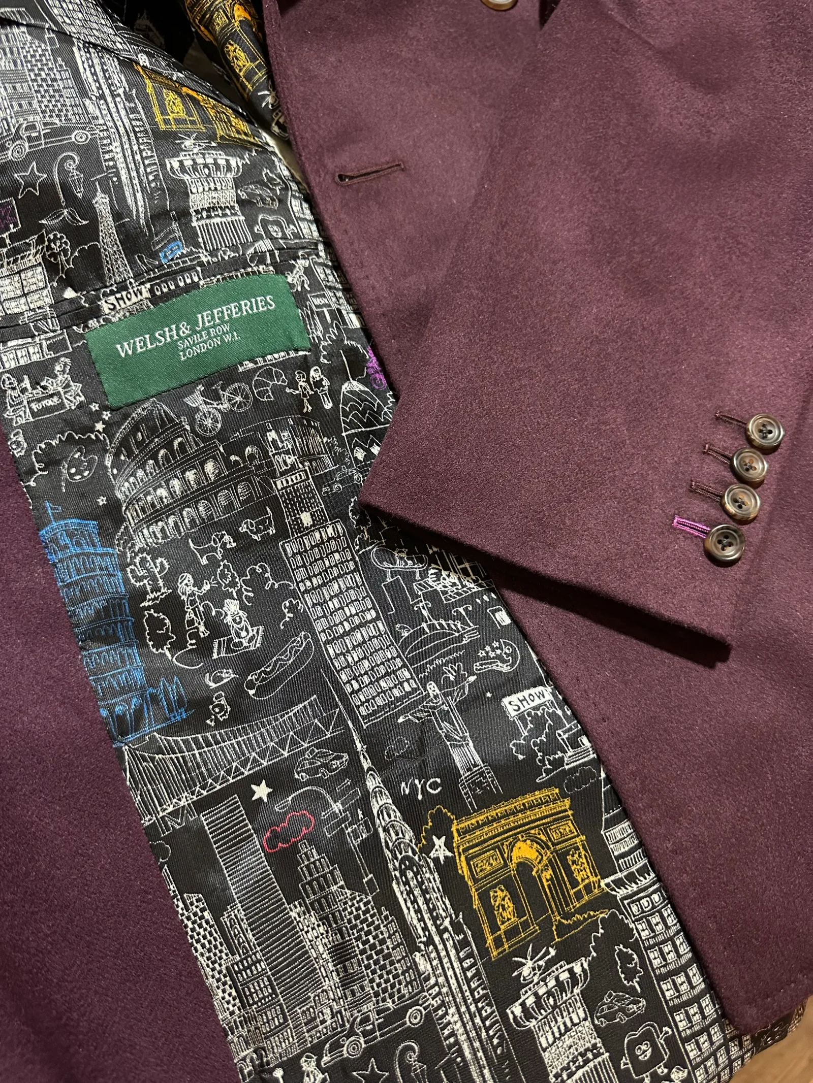Interior view of a deep mulberry double-breasted jacket, revealing a black-and-white illustrated London and New York city-print lining.