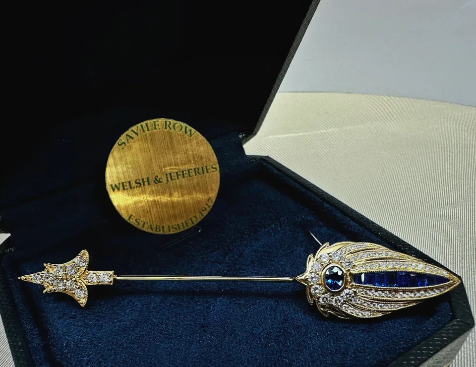 Gold feather stick pin with blue sapphires, diamond-set ribs and a fleur-de-lis guard displayed in a navy case.
