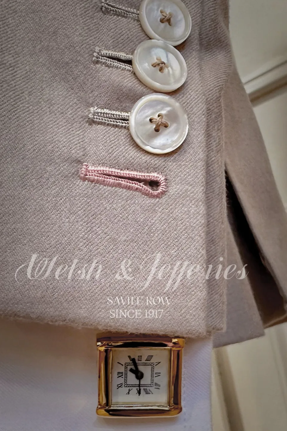Small square gold dress watch peeking beneath a pale tailored cuff with mother-of-pearl buttons and a pink buttonhole.