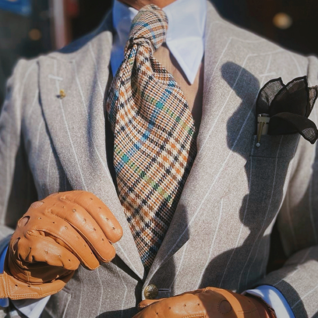 Crowned gold cypher lapel pin in a sunlit tailoring portrait with a generous checked tie and caramel driving gloves.