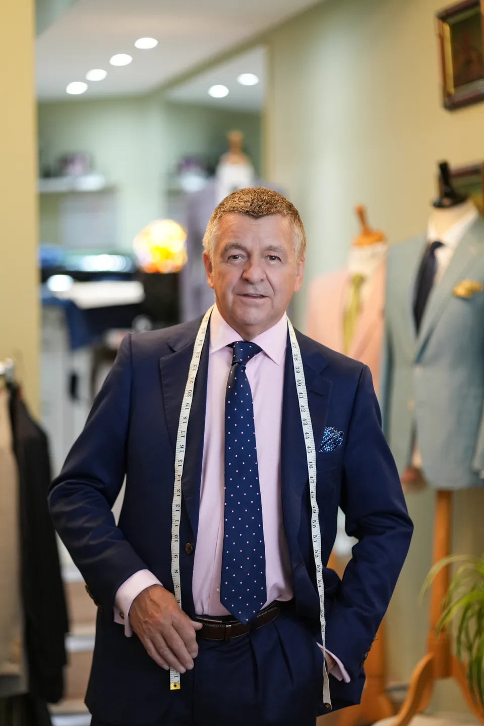 A senior tailor stands in a navy suit with measuring tape at the ready.