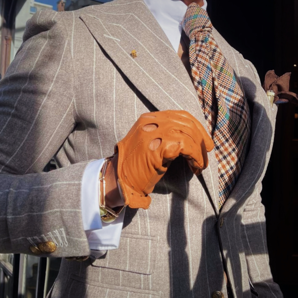 Tiny crowned gold cypher on the lapel of a pale pinstriped suit, framed by a checked tie and tan driving glove.