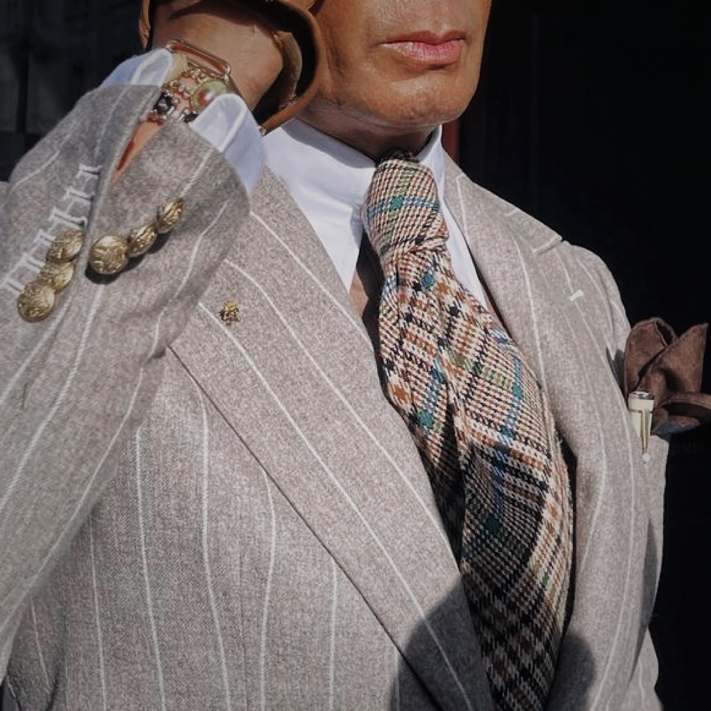 Gold crowned cypher pin on a grey pinstriped lapel beside sculpted cuff buttons, a patterned tie and a dress watch.